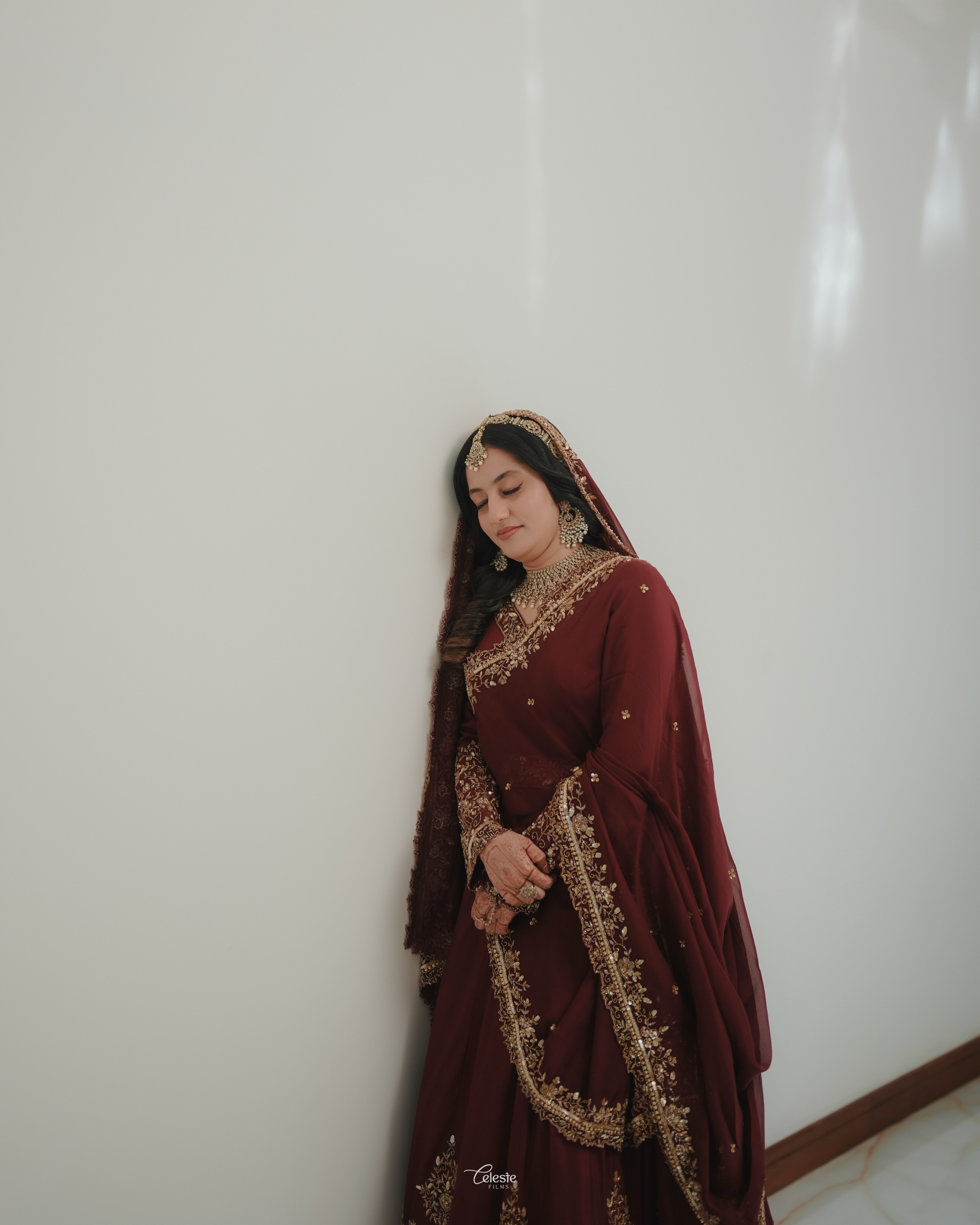 Bride in maroon wedding attire standing against a light wall
